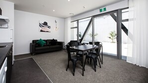 Een moderne kamer met een eettafel, stoelen, een zwarte bank met een groen kussen, een schilderij en een groot raam met uitzicht op palmbomen.