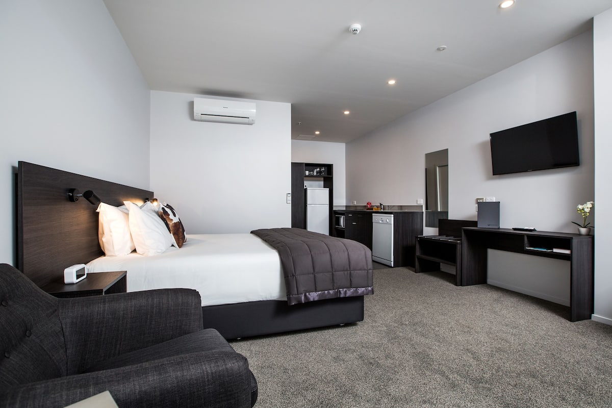 A modern hotel room with a bed, a desk, a TV, and a kitchenette.