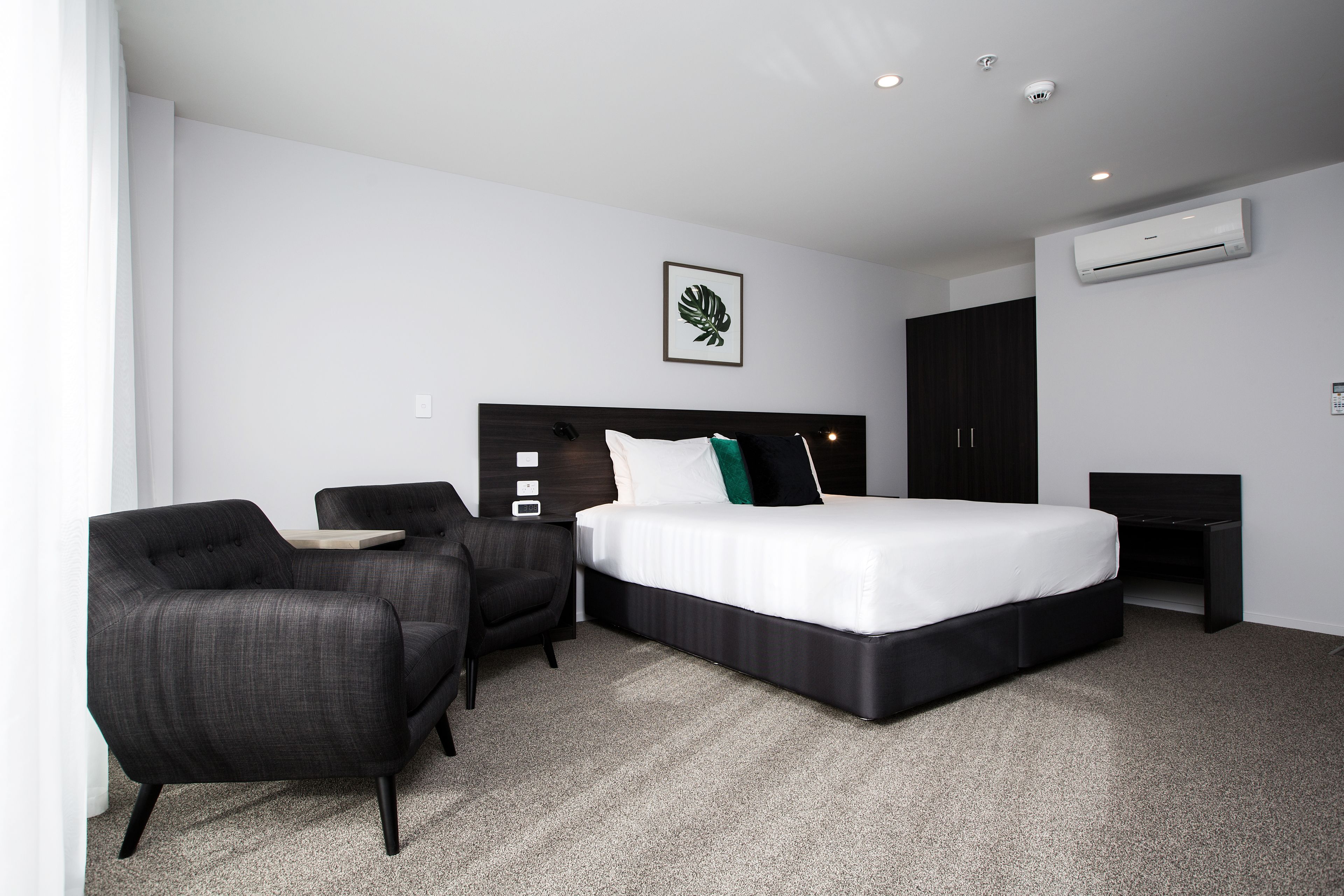 A modern hotel room with a large bed, a desk, a chair, and a wall-mounted air conditioner.