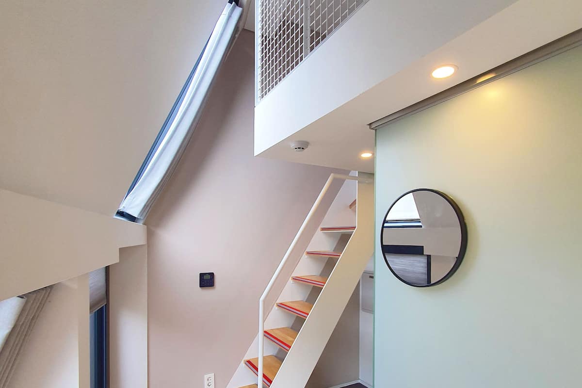 Un interior moderno con una escalera de madera, un espejo redondo y una rejilla de ventilación en el techo.