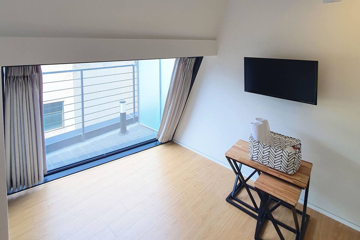 Una habitación con un ventanal grande, un televisor de pantalla plana en la pared y una pequeña mesa de madera con un cojín estampado.