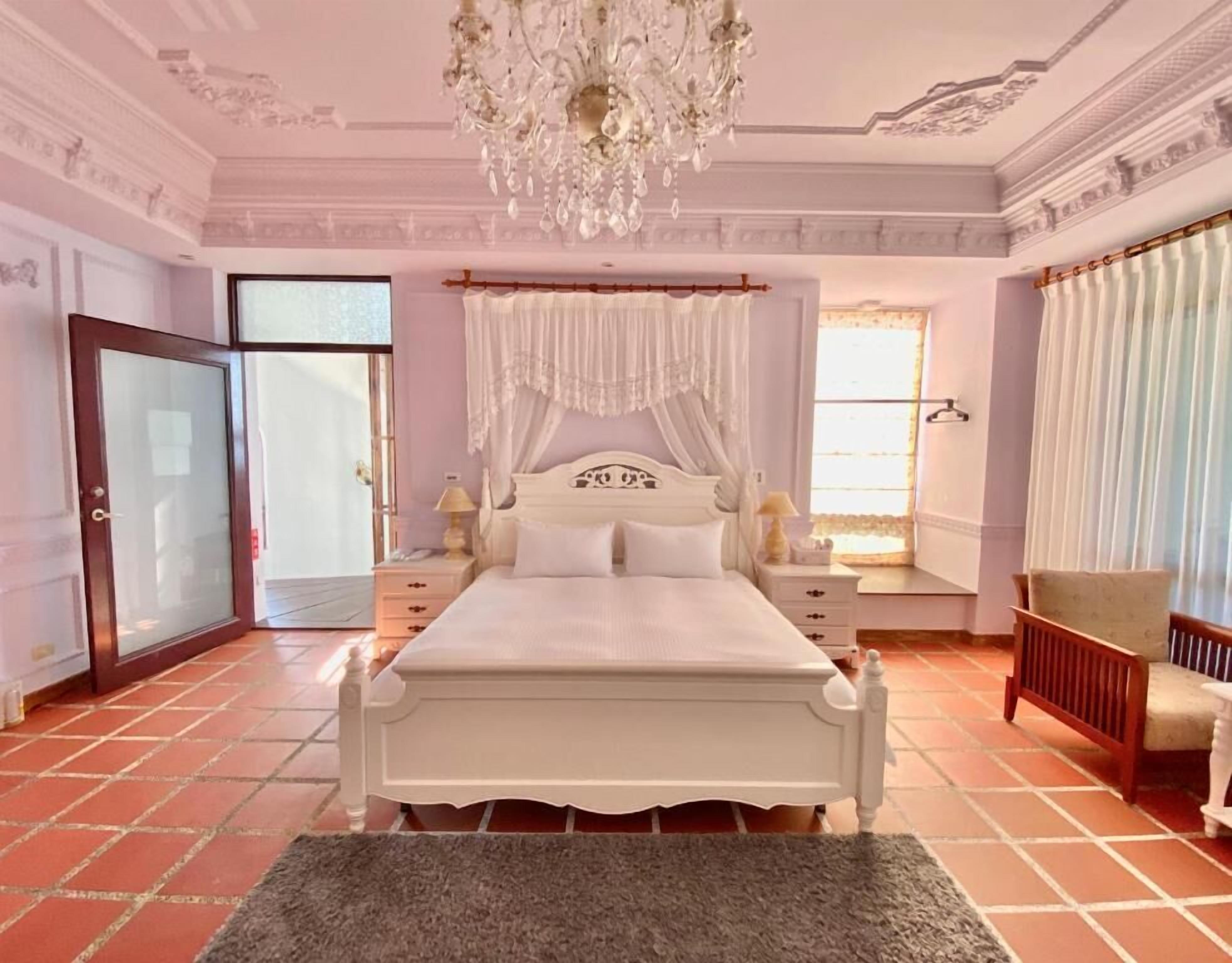 A bedroom with a white bed, a chandelier, a window with curtains, a chair, and a door.