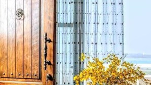 A wooden door with a brass handle, a table with a vase of yellow flowers, a glass of orange juice, and a view of the sea through a window with sheer curtains. - 23.5 Blue B & B (Xiyu)
