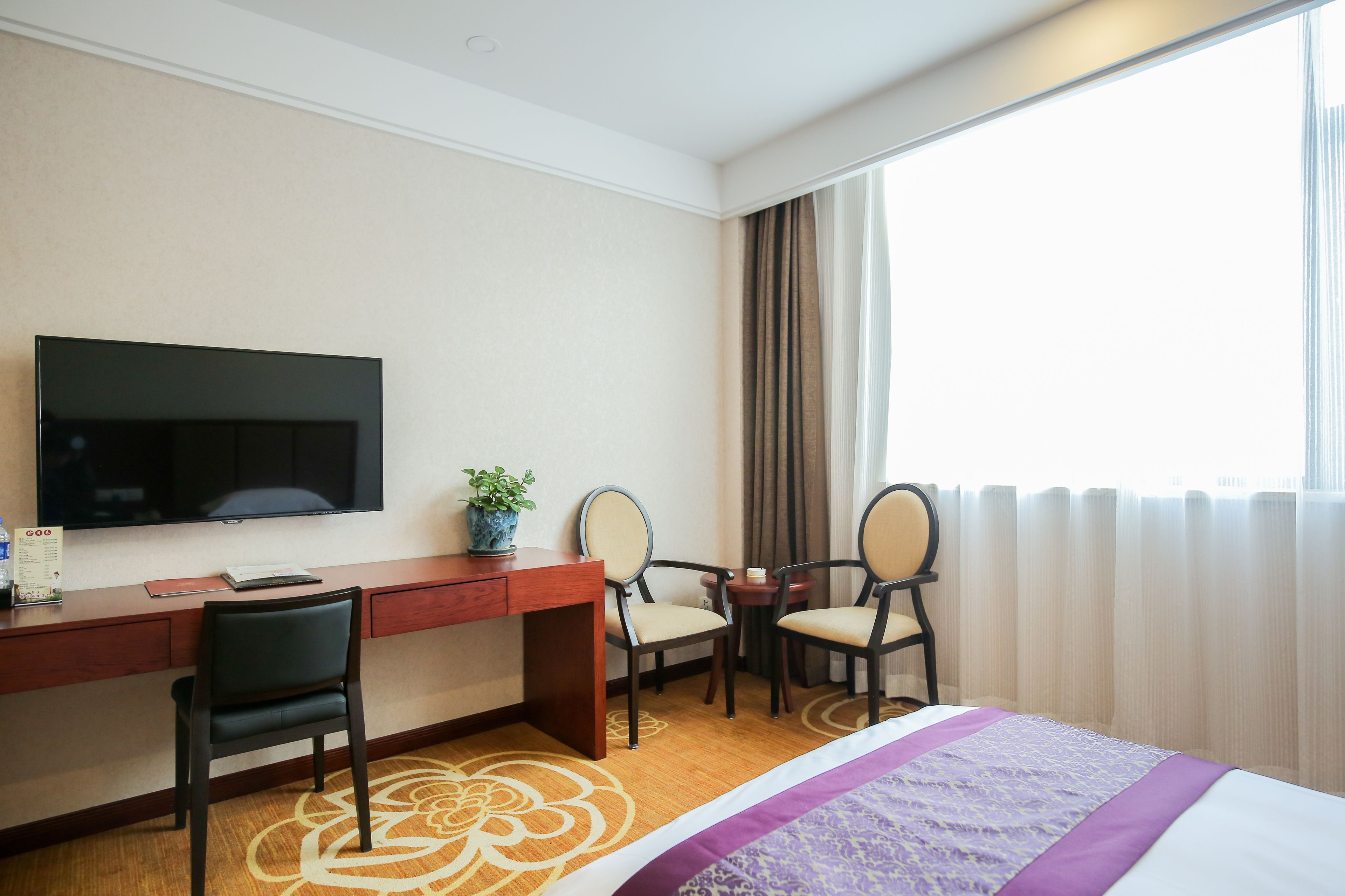 A hotel room with a bed, a desk, a television, two chairs, a small table, and a window with curtains.
