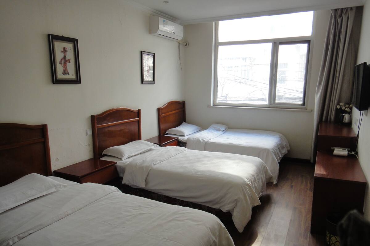 Une chambre d’hôtel avec trois lits simples, une table de chevet en bois, un petit bureau avec une chaise, une fenêtre donnant sur des immeubles et un climatiseur fixé au mur.