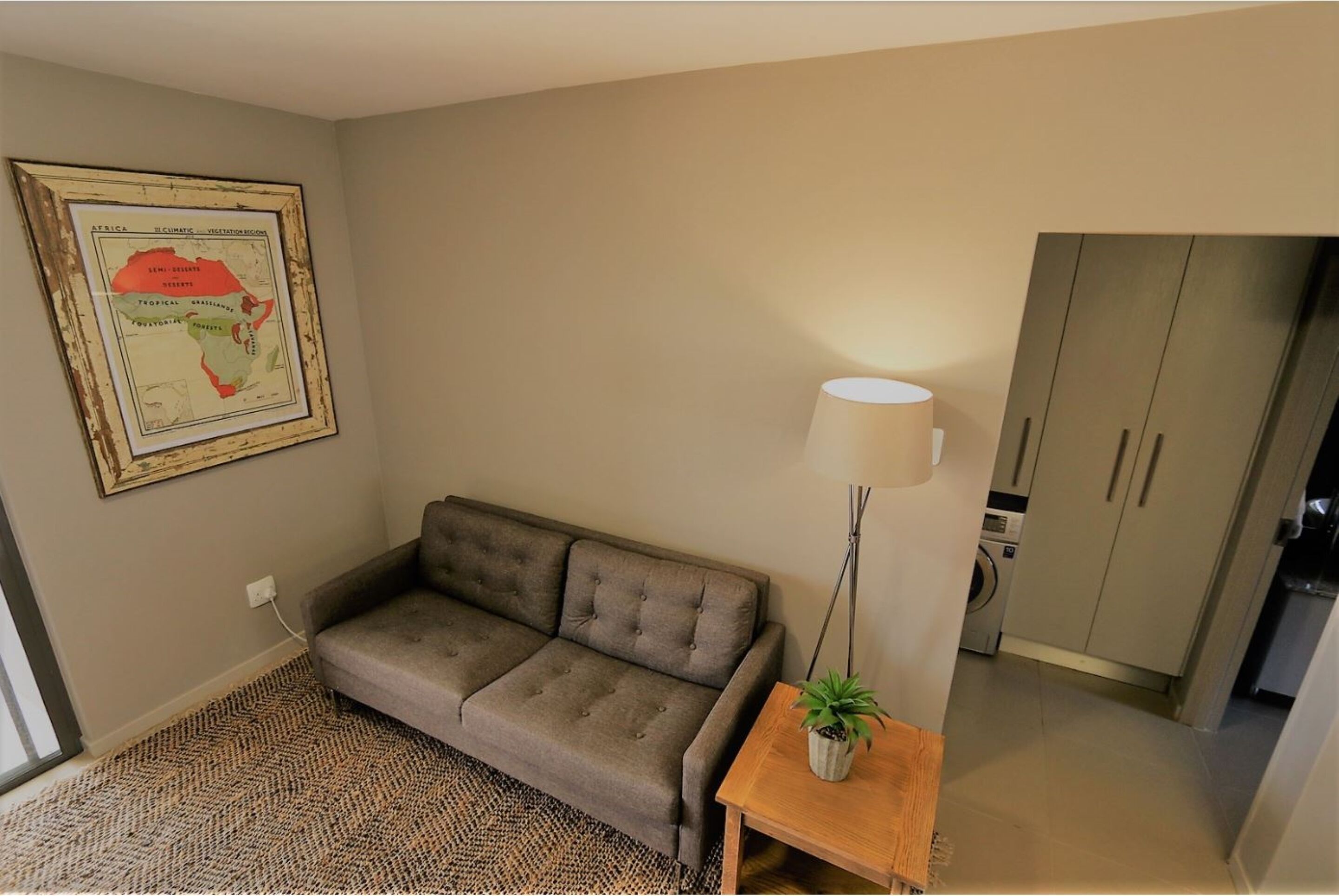 A living room with a sofa, a lamp, a small table with a plant, and a framed map on the wall.