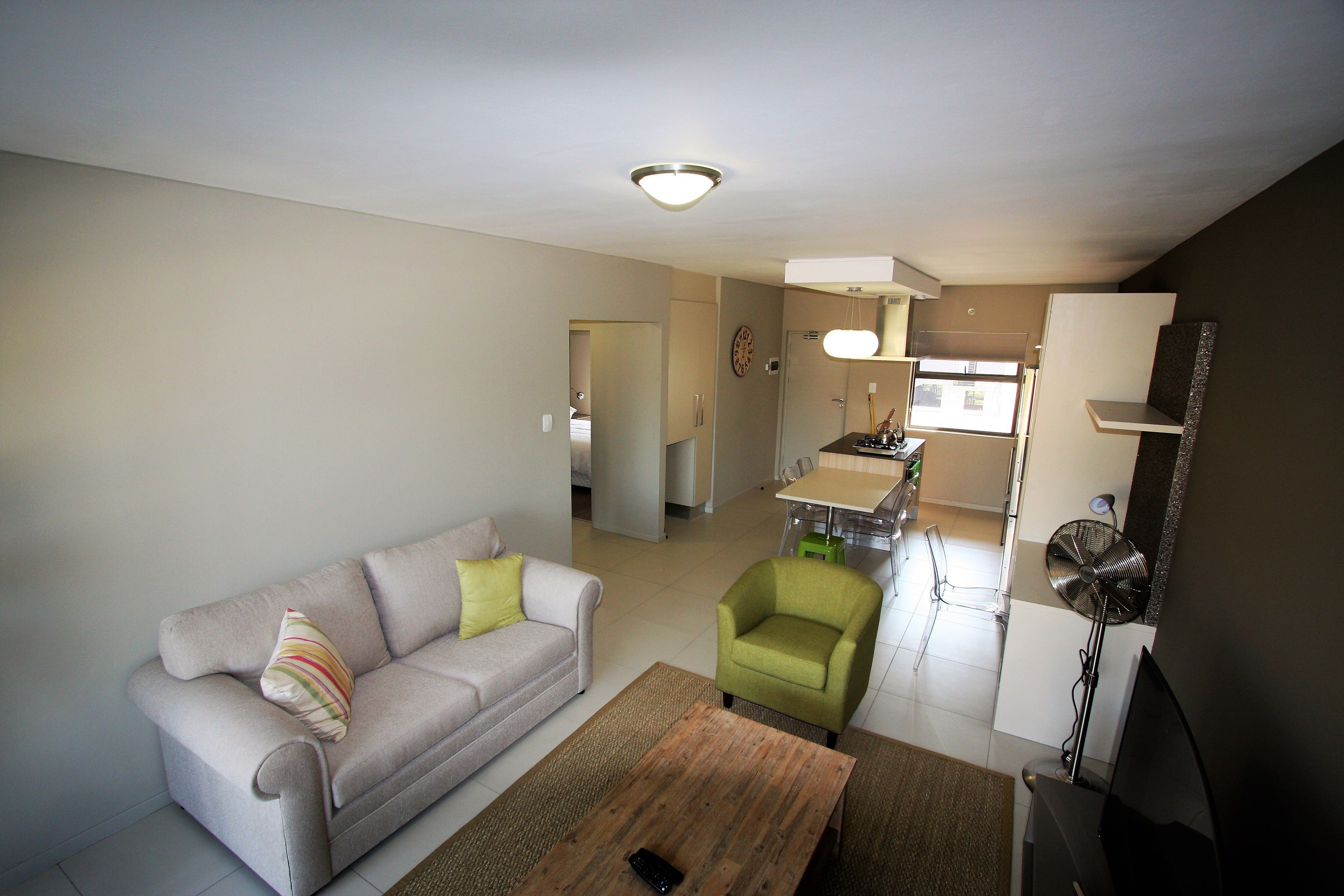A living room with a sofa, a green armchair, and a coffee table. There is a kitchen in the background with a window and a fan.