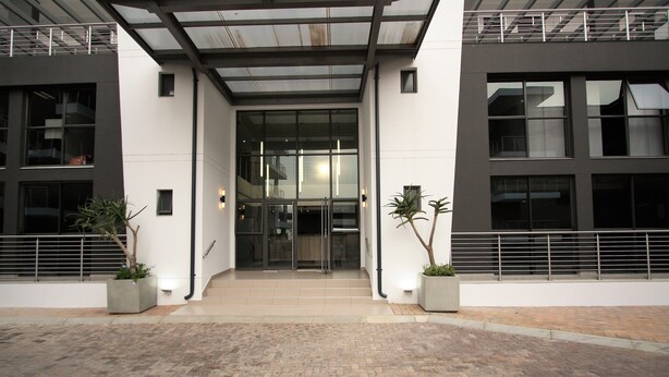 A modern building entrance with a glass door, potted plants, and a tiled pavement.