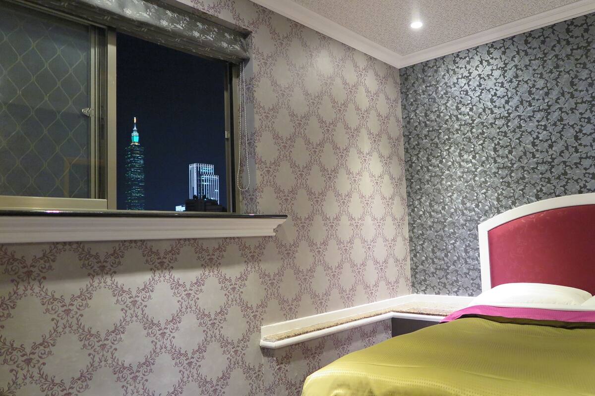 Een slaapkamer met een bed, een raam met uitzicht op een stadsbeeld, behang met een patroon en een rode hoofdeinde.
