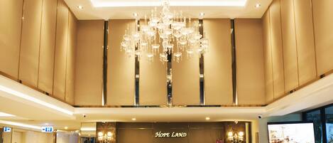 A hotel lobby with a marble reception desk, a large chandelier, and a flat-screen TV on the wall.