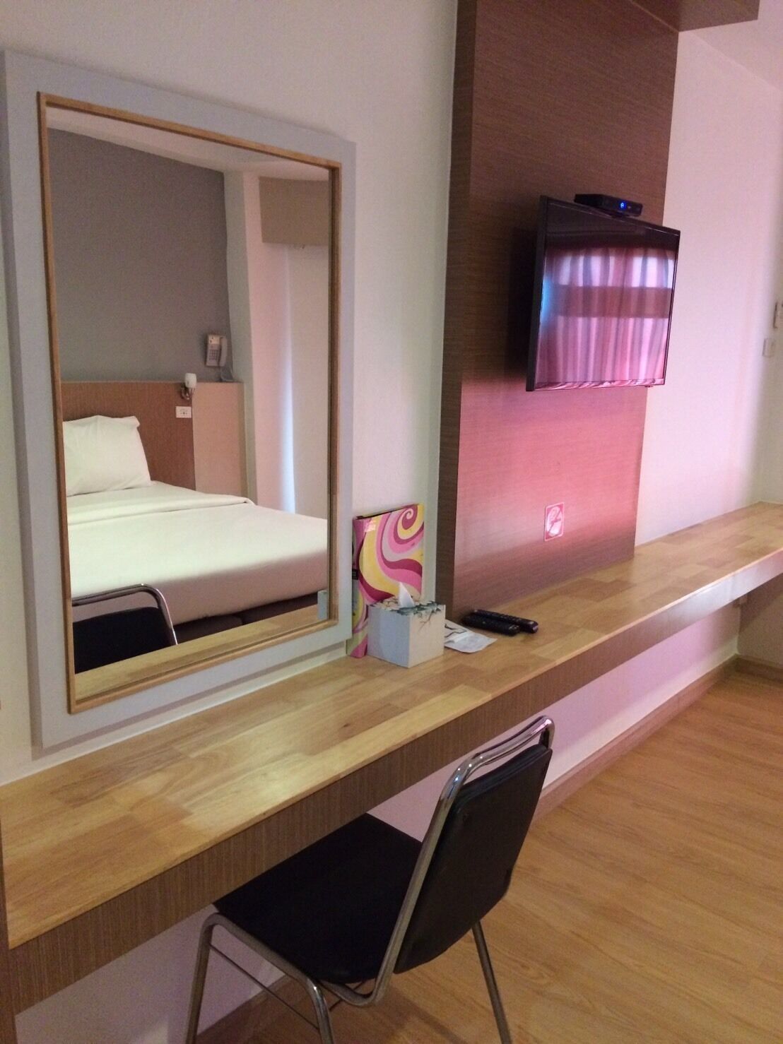 A hotel room with a bed, a desk with a chair, a TV mounted on the wall, and a mirror above the desk.