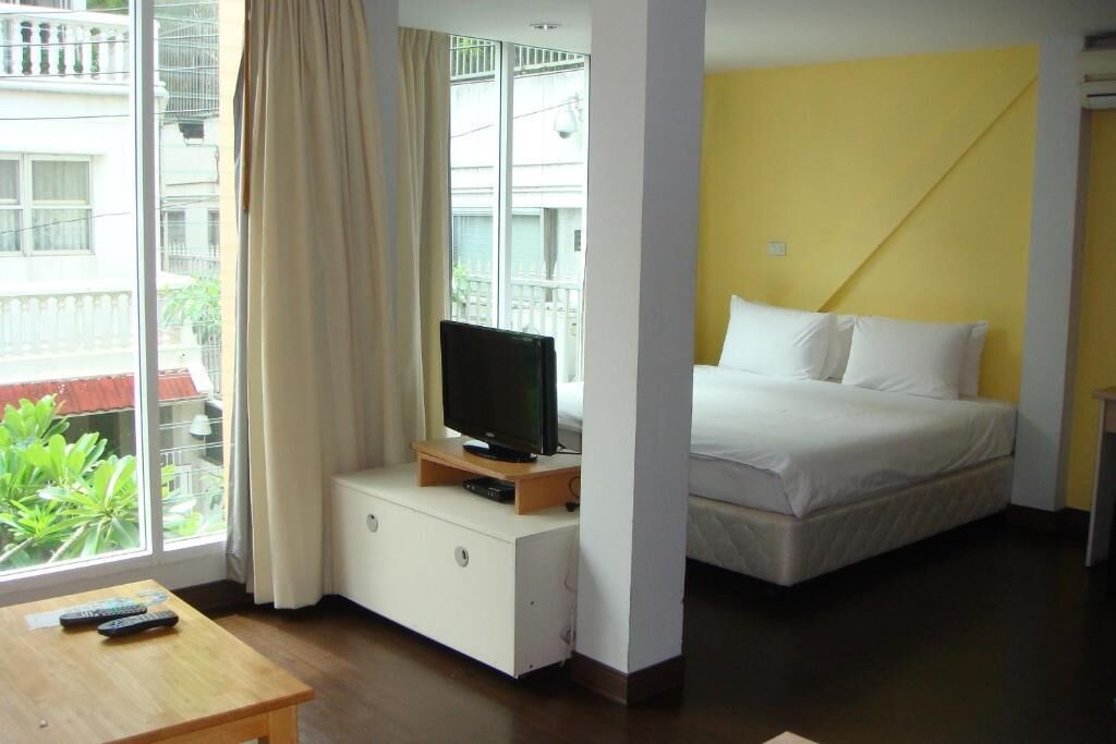 Une chambre d’hôtel avec un lit, un téléviseur posé sur un meuble en bois, une table avec des télécommandes et une vue sur un balcon agrémenté de verdure.