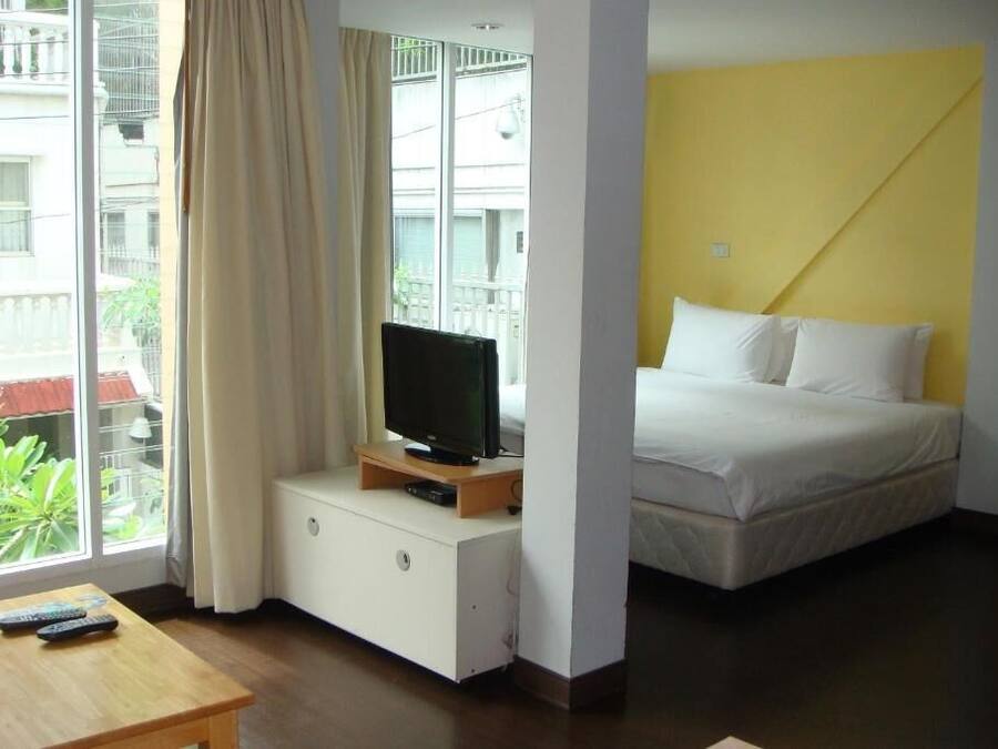 Une chambre d’hôtel avec un lit, un téléviseur posé sur un meuble en bois, une table avec des télécommandes et une vue sur un balcon agrémenté de verdure.