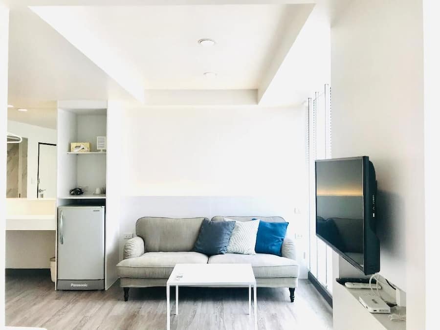 A modern living room with a sofa, a flat-screen TV, a white coffee table, and a built-in kitchen unit.