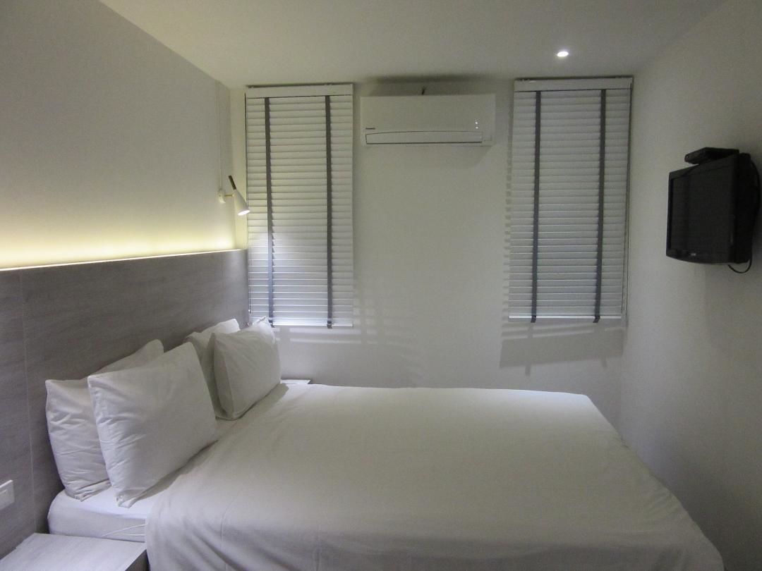 A bedroom with a bed, two windows with blinds, an air conditioning unit, and a television mounted on the wall.