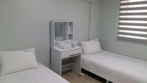 A small, clean room with two single beds, a white dresser with a mirror and a basket, and a window with blinds. - MK Guest House (Busan)