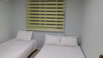 Une chambre avec deux lits simples, chacun doté d’un matelas blanc et d’oreillers assortis, et une fenêtre avec des stores horizontaux.