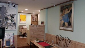A room with a wooden bench, a table, and a TV. There are paintings on the walls and a window with blinds. - MK Guest House (Busan)