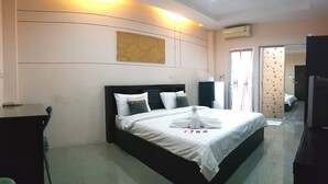 A hotel room with a large bed, a desk, and a chair. There is a window with curtains, an air conditioning unit on the wall, and a door leading to another room.