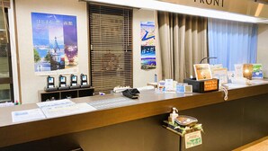 A hotel reception desk with a 'FRONT' sign, a window with blinds, and various informational posters and signs. - A-Gate Hotel (Hakodate)