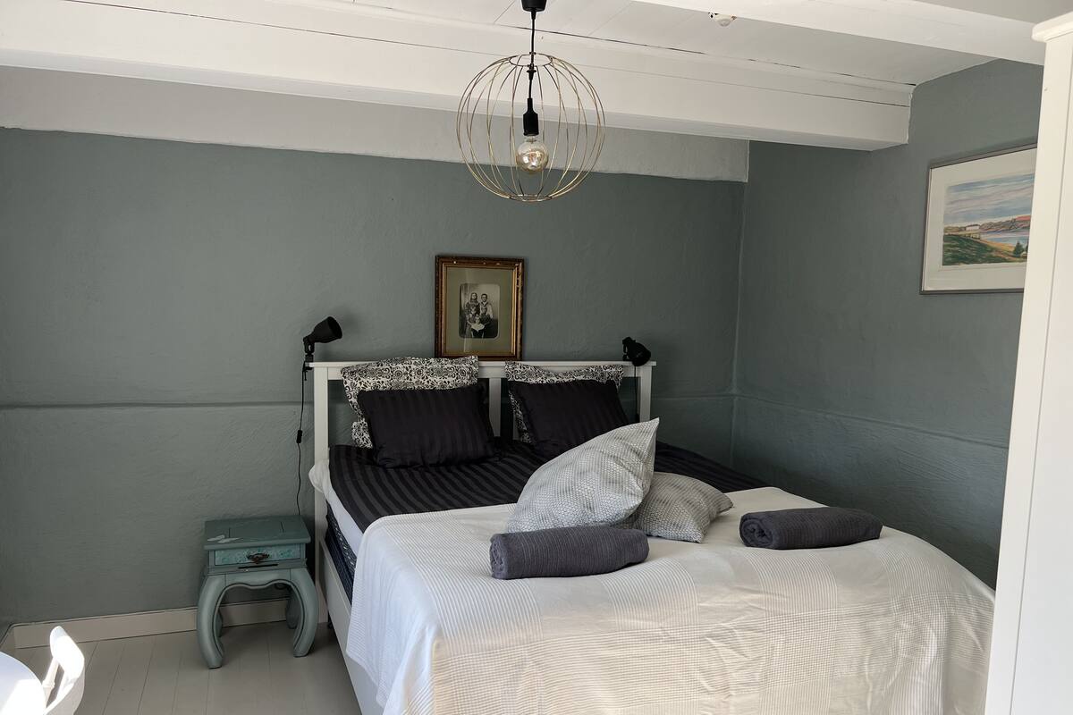A bedroom with a white bed, grey walls, a small bedside table, a lamp, and a framed picture on the wall.