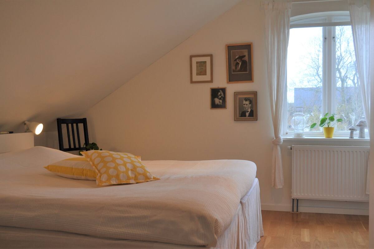 A bedroom with a bed, a nightstand, a lamp, a window with curtains, and wall-mounted pictures.