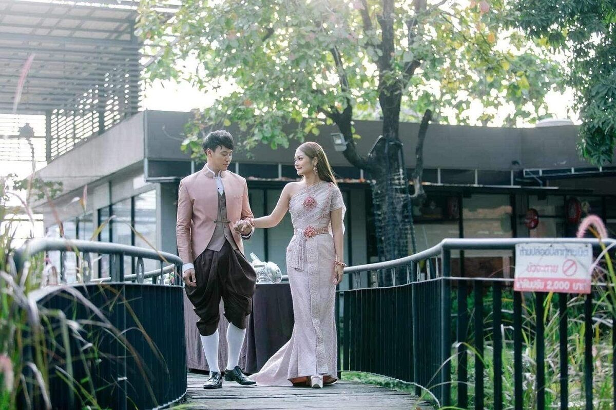 Una pareja tomándose de las manos sobre un puente, con un edificio y follaje al fondo.
