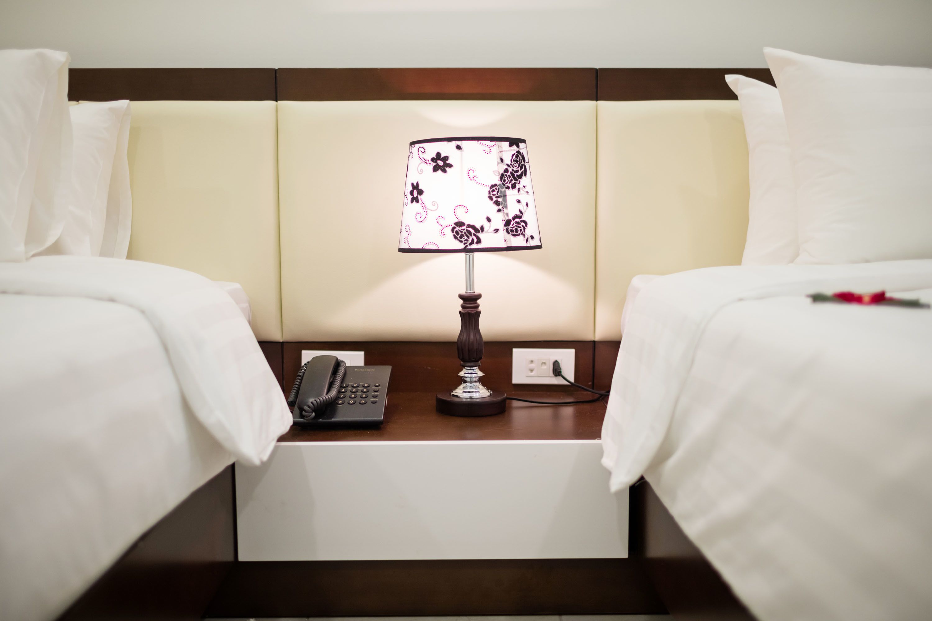 A hotel room with two beds, a nightstand with a lamp, a phone, and a power outlet.