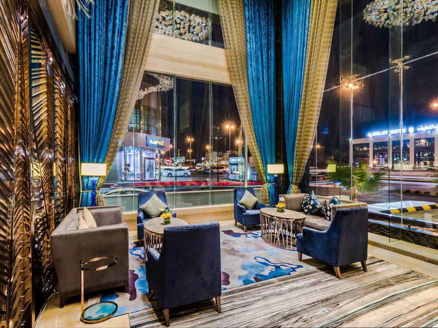 A hotel lobby with a view of the street, featuring a seating area with sofas and a coffee table, large windows with curtains, and a decorative wall.