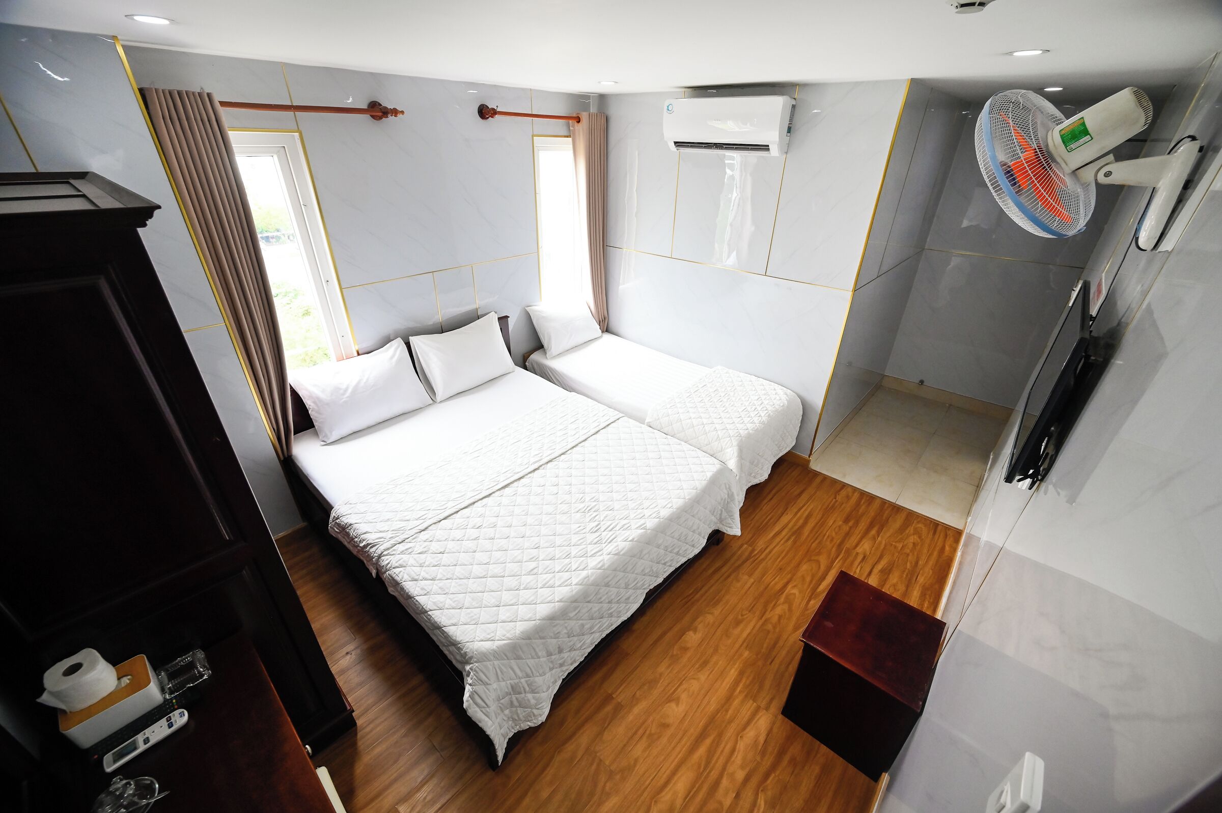 A hotel room with a bed, a wooden floor, a wardrobe, an air conditioning unit, and a ceiling fan.
