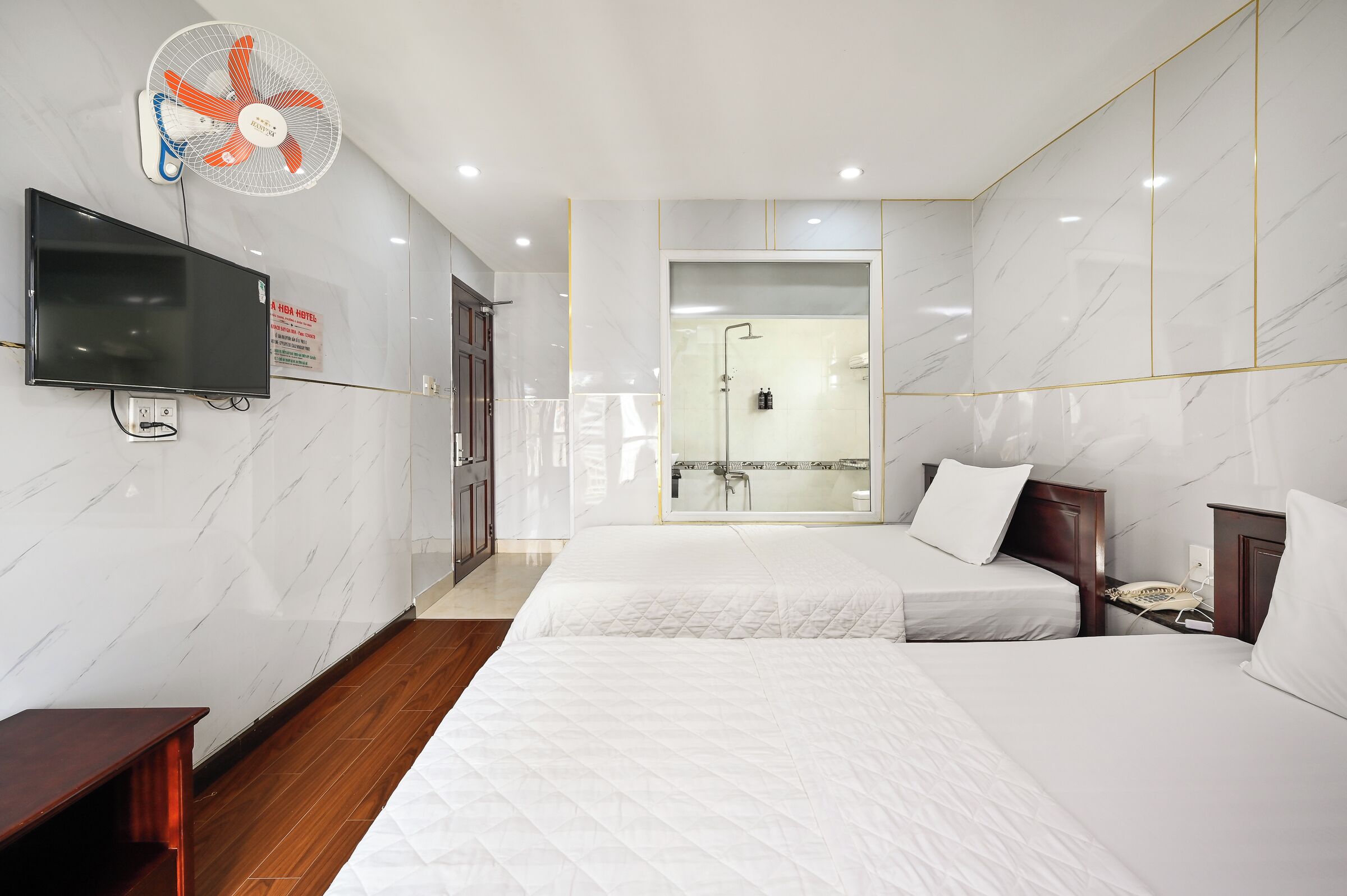 A hotel room with two beds, a TV, a fan, a shower, and a sign indicating it's a Hida Hotel.