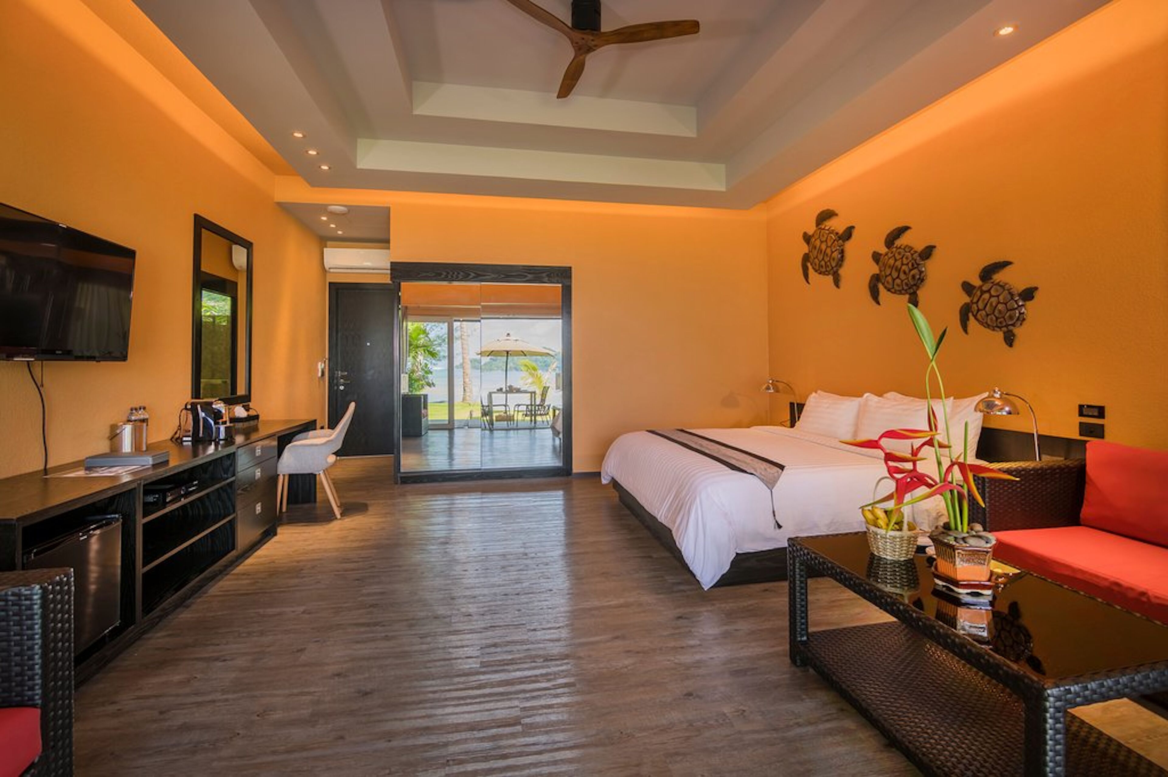 A hotel room with a bed, a desk, a TV, and a balcony with a view of palm trees.