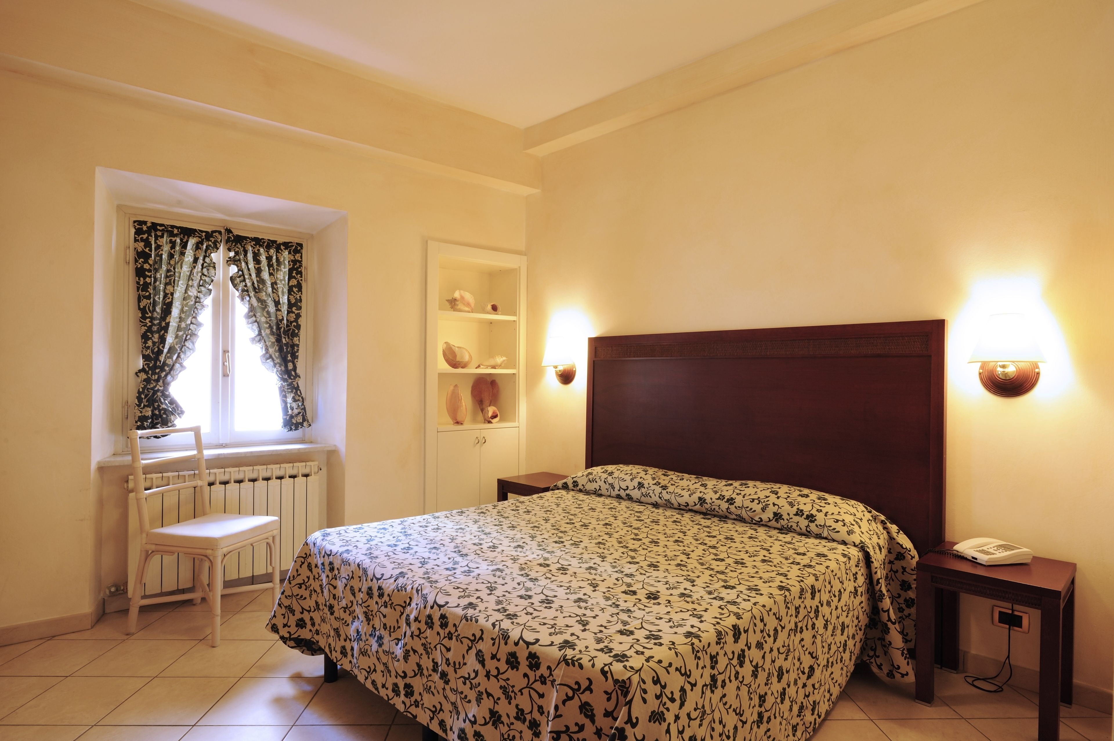 A bedroom with a bed, a chair, a shelf with decorative items, and a bedside table with a lamp.