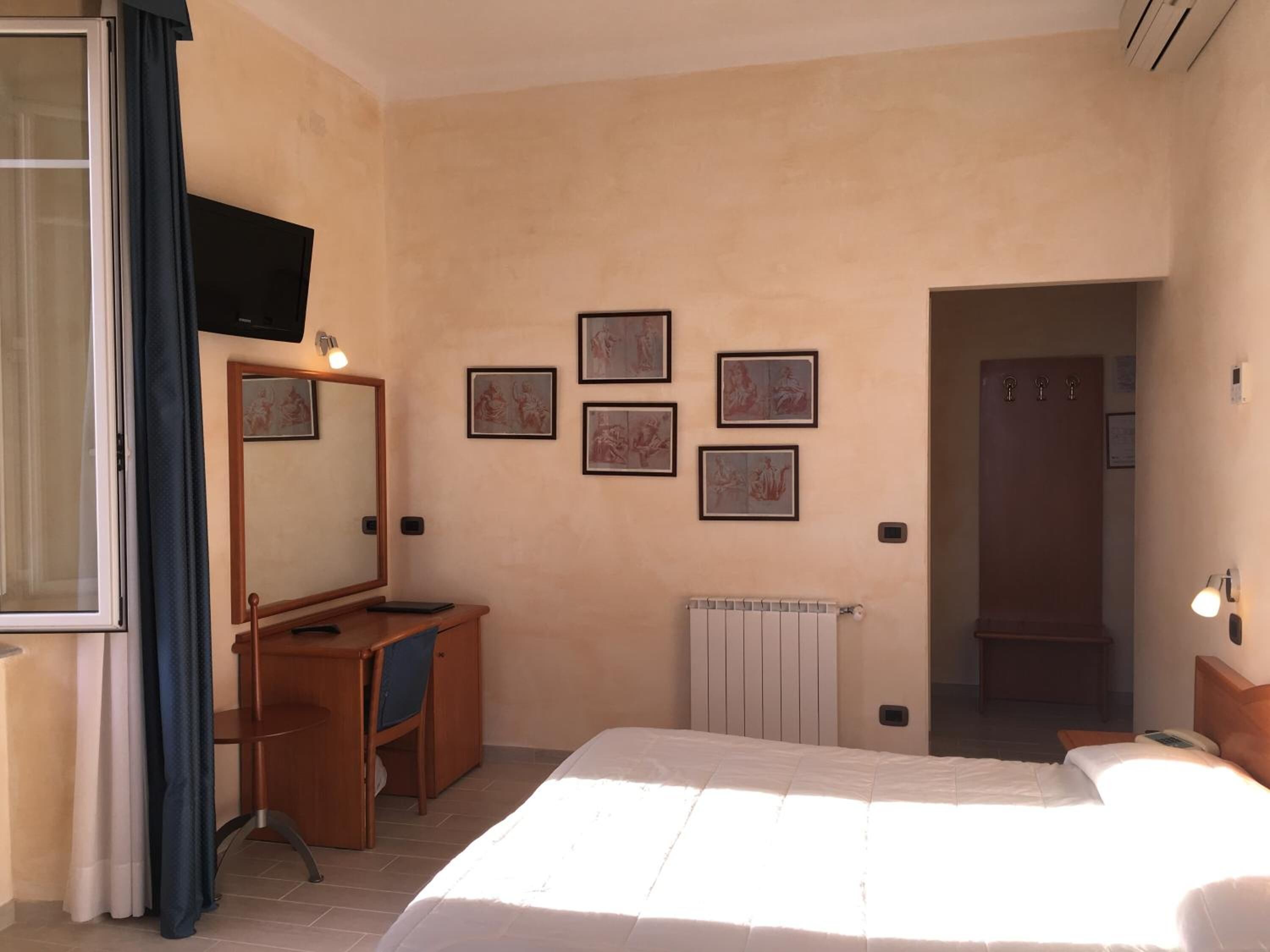 A hotel room with a bed, a desk with a chair, a mirror, a television, and framed artwork on the wall.
