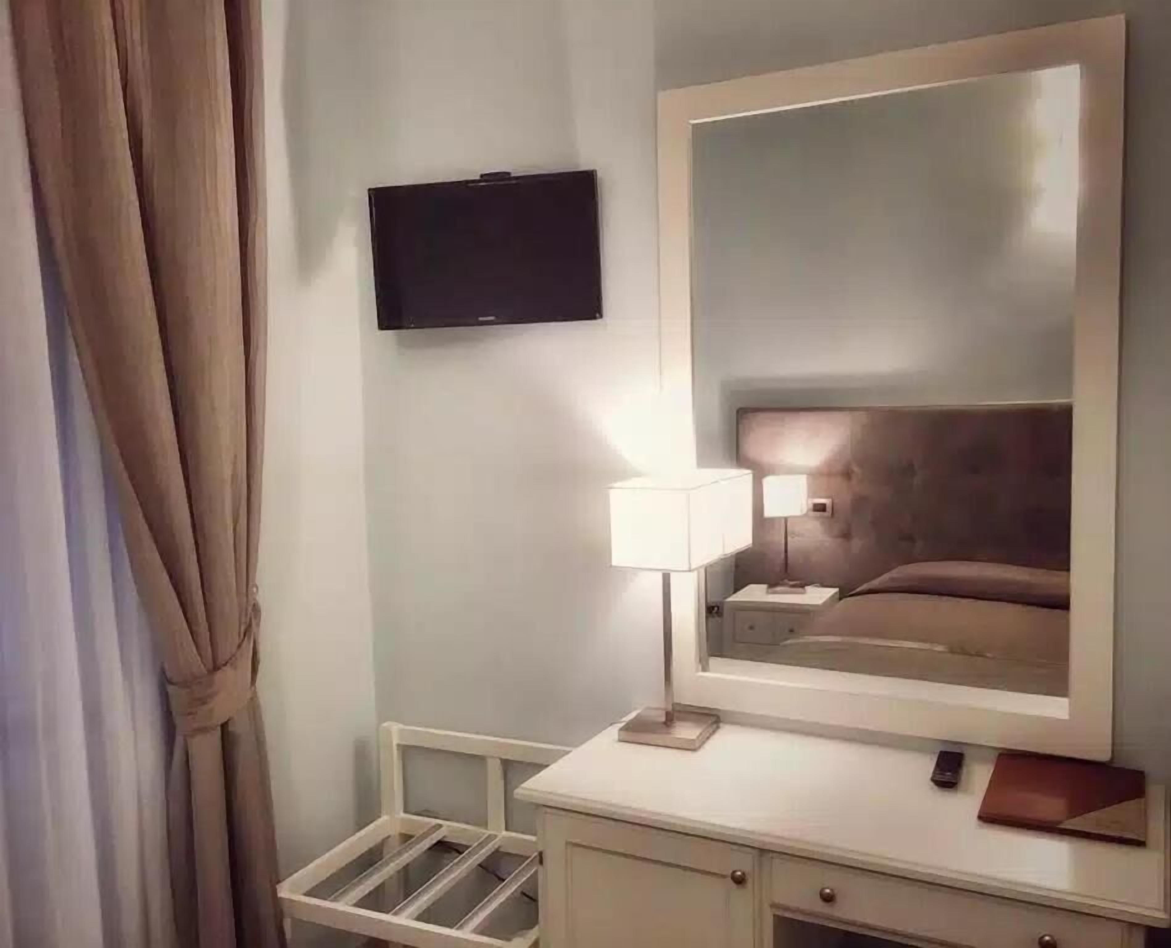 A hotel room with a large mirror, a flat-screen TV mounted on the wall, a bedside table with a lamp, and a small drawer unit.