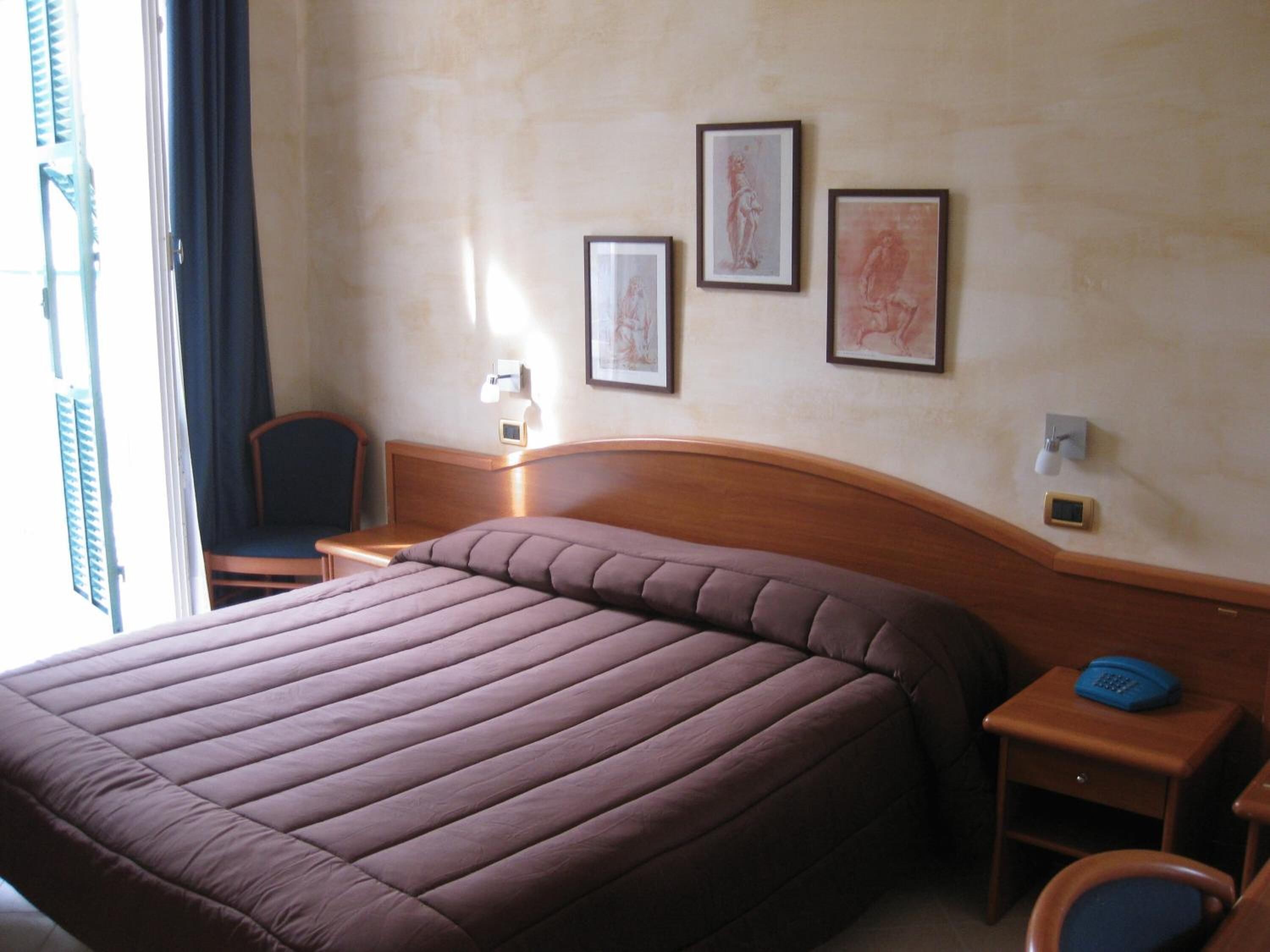 Una habitación de hotel con una cama grande, una mesita de noche con lámpara, una silla y una obra de arte en la pared.