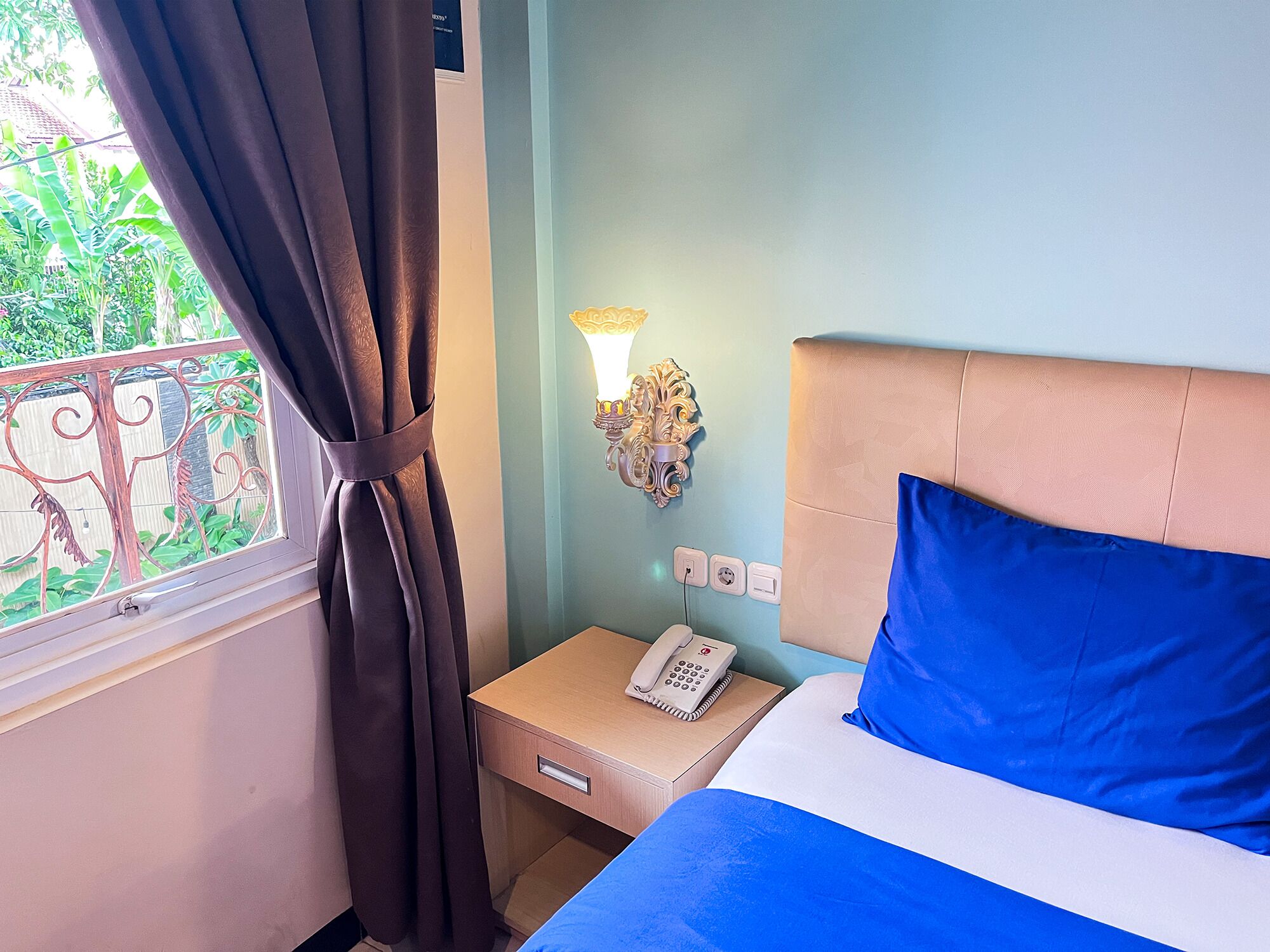Ein Hotelzimmer mit einem Bett, einem Nachttisch mit Telefon, einer Wandleuchte und einem Fenster mit Vorhängen.