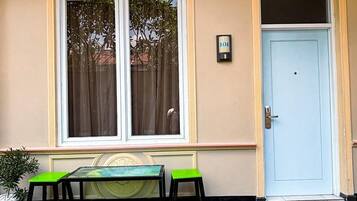 A building facade with a beige wall, a blue door, and a window with a curtain. There is a green bench and a small table in front.