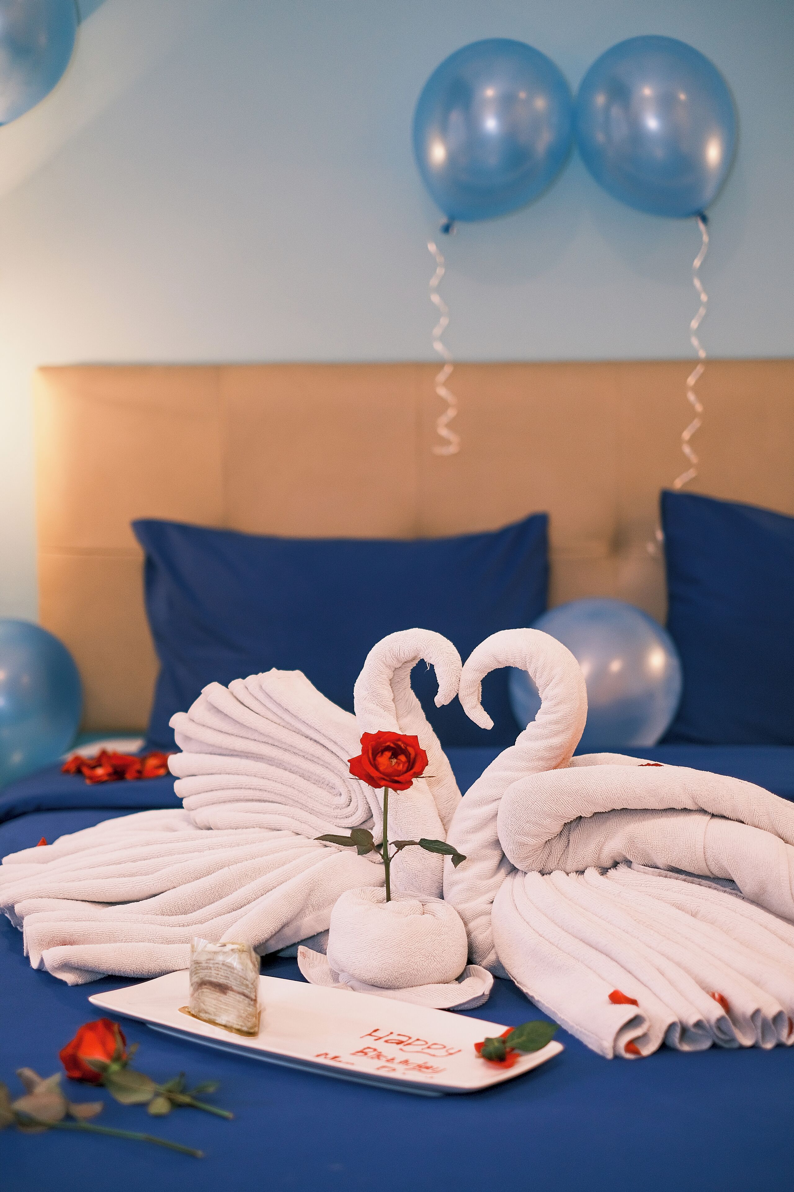 Een netjes opgemaakt bed met blauwe kussens, een boeket rode rozen en een zwaan-vormige handdoekopstelling op een witte schaal.