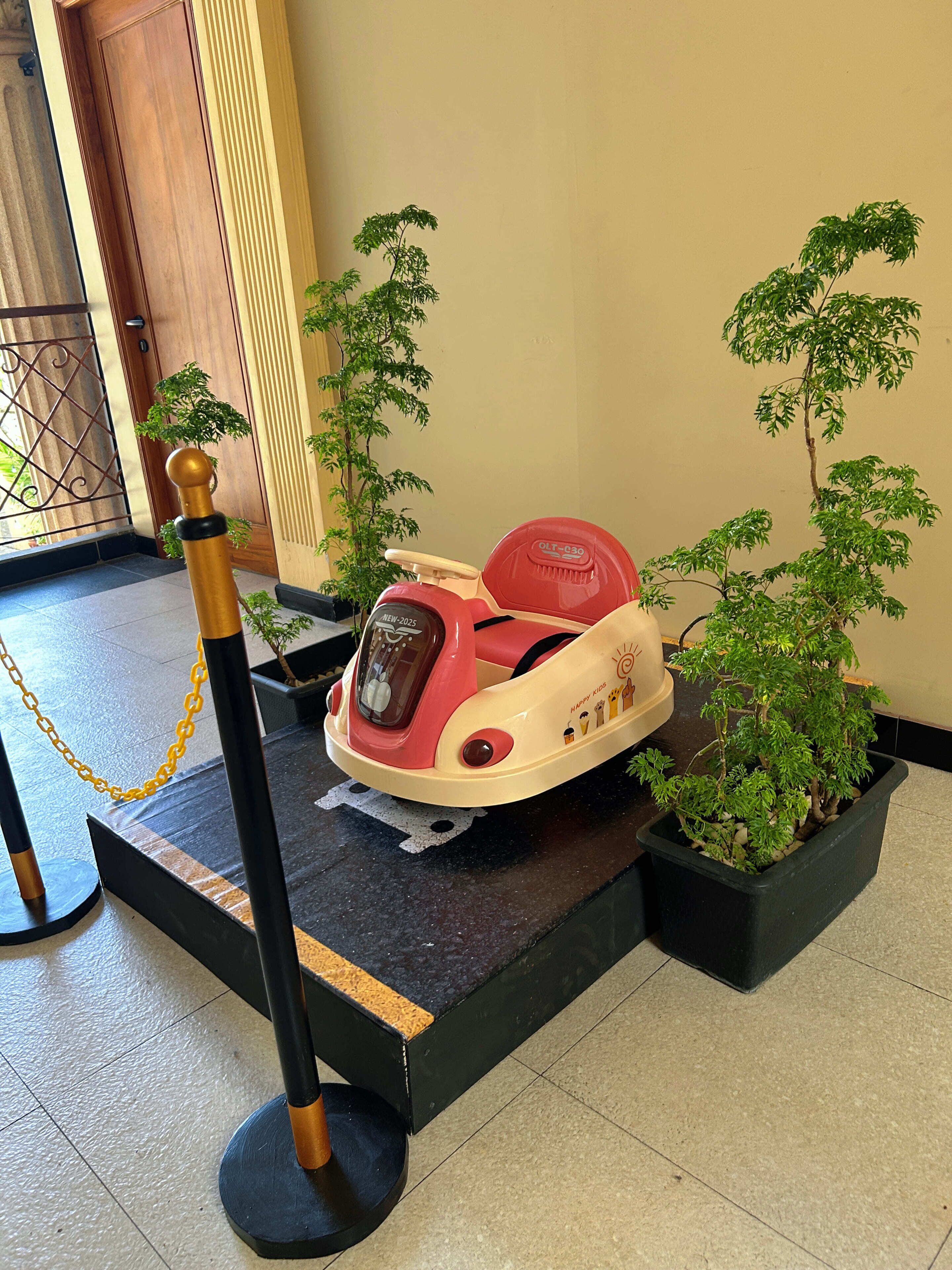 A red and white ride-on toy car with a black base, surrounded by potted plants and a yellow chain barrier.