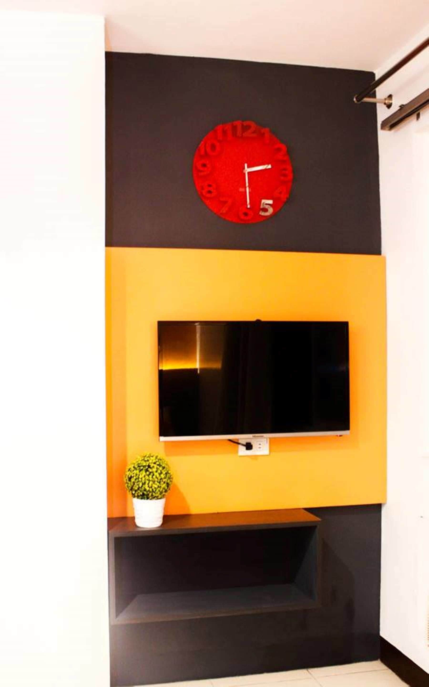 A room with a black and yellow wall, a red clock, a flat-screen TV, and a small potted plant.