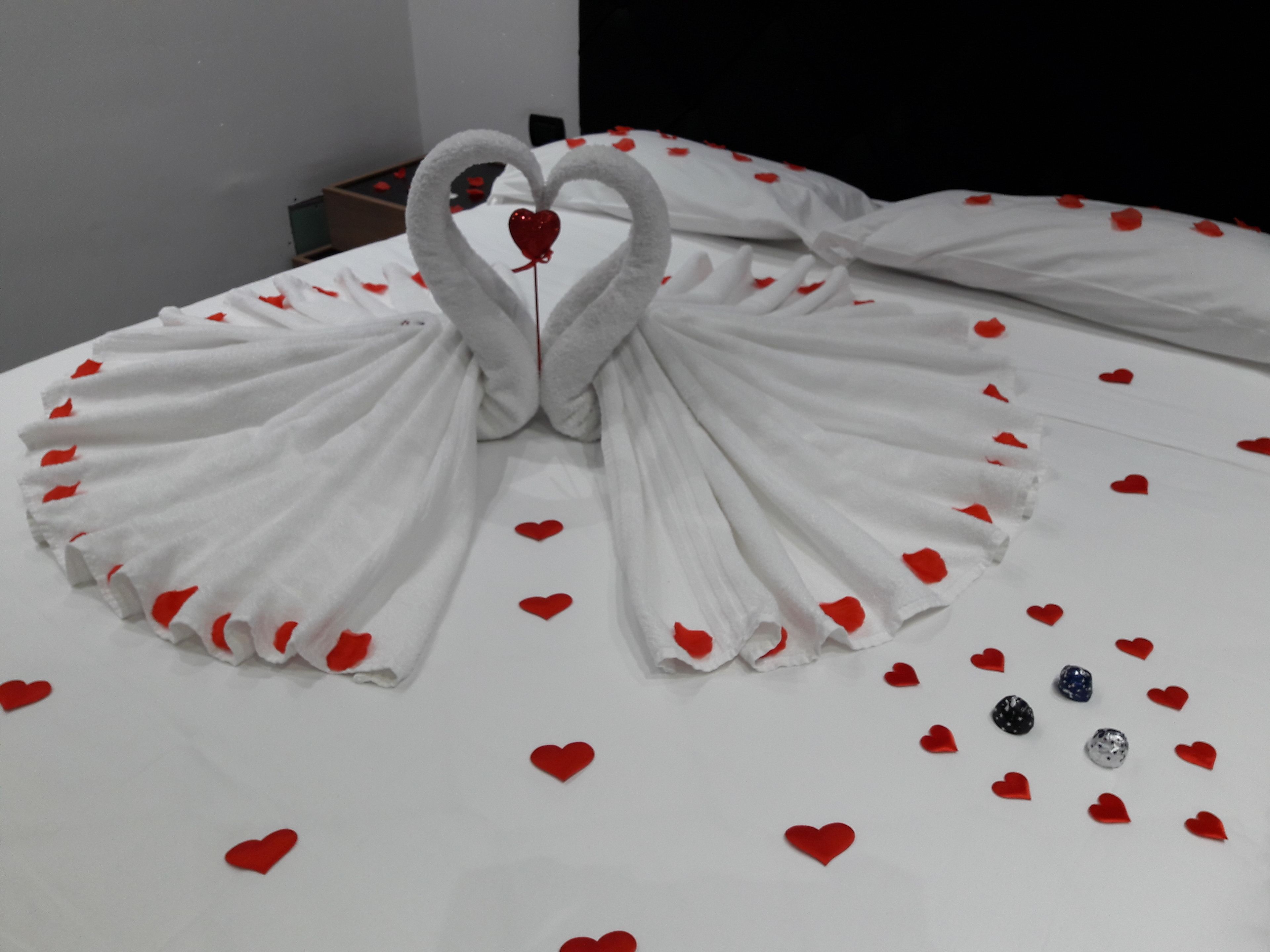 A bed with a white bedspread and red heart decorations, including a heart-shaped towel arrangement and scattered heart-shaped confetti.