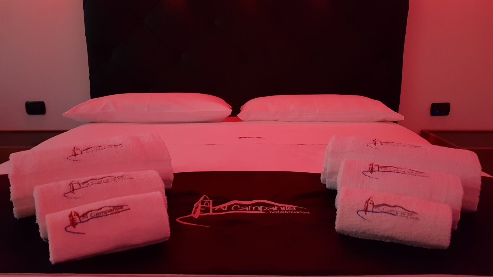 Una cama con sábanas y almohadas blancas, un logotipo en la colcha y toallas con un logotipo sobre una mesita de noche.