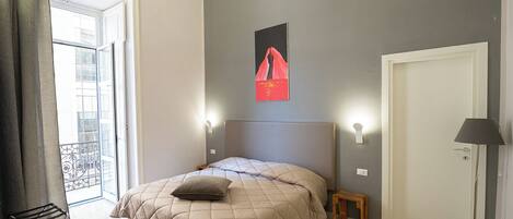 A bedroom with a bed, a small bedside table, a lamp, a door, and a window with a view of buildings.