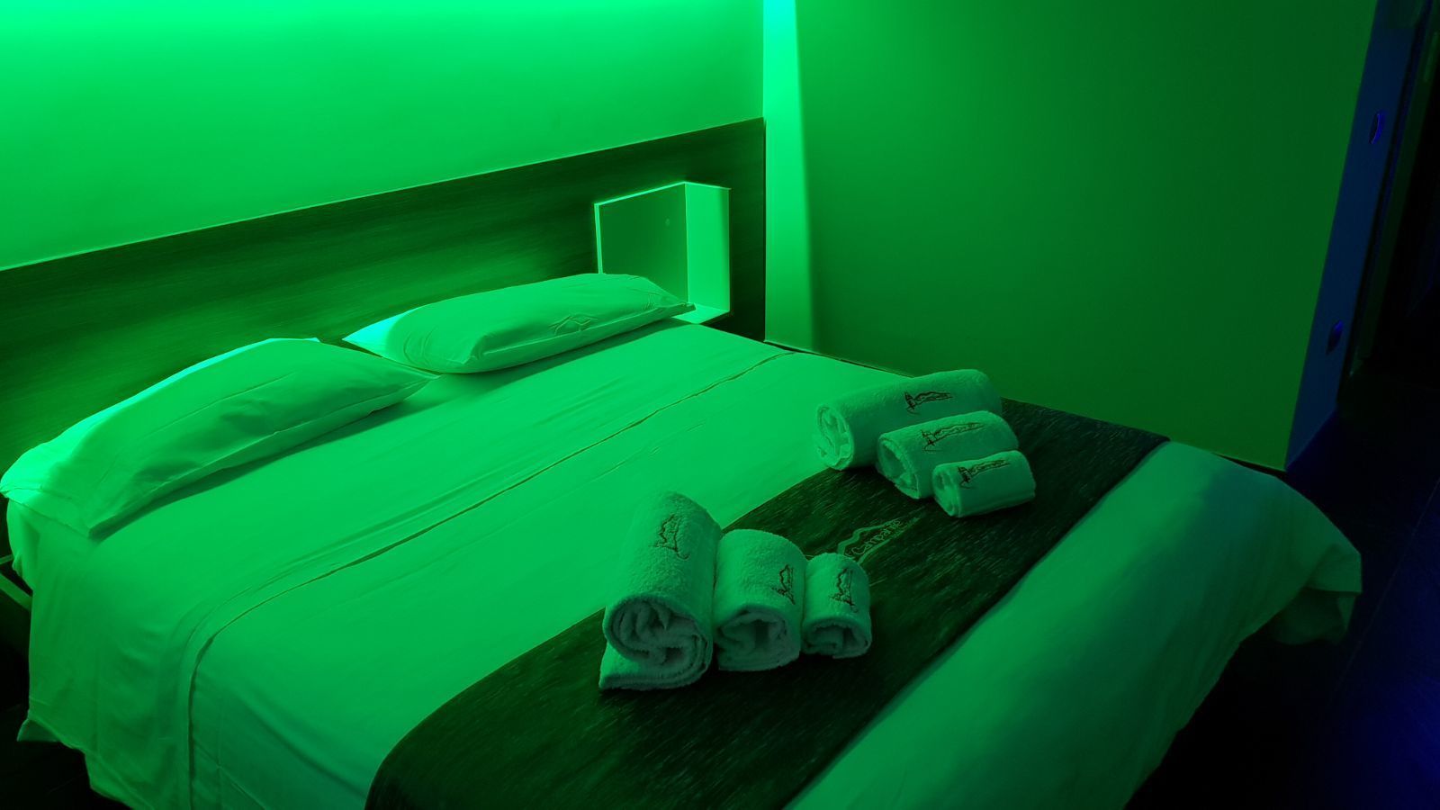 A bed with a headboard, a wooden bedside table with rolled towels, and a green ambient light.
