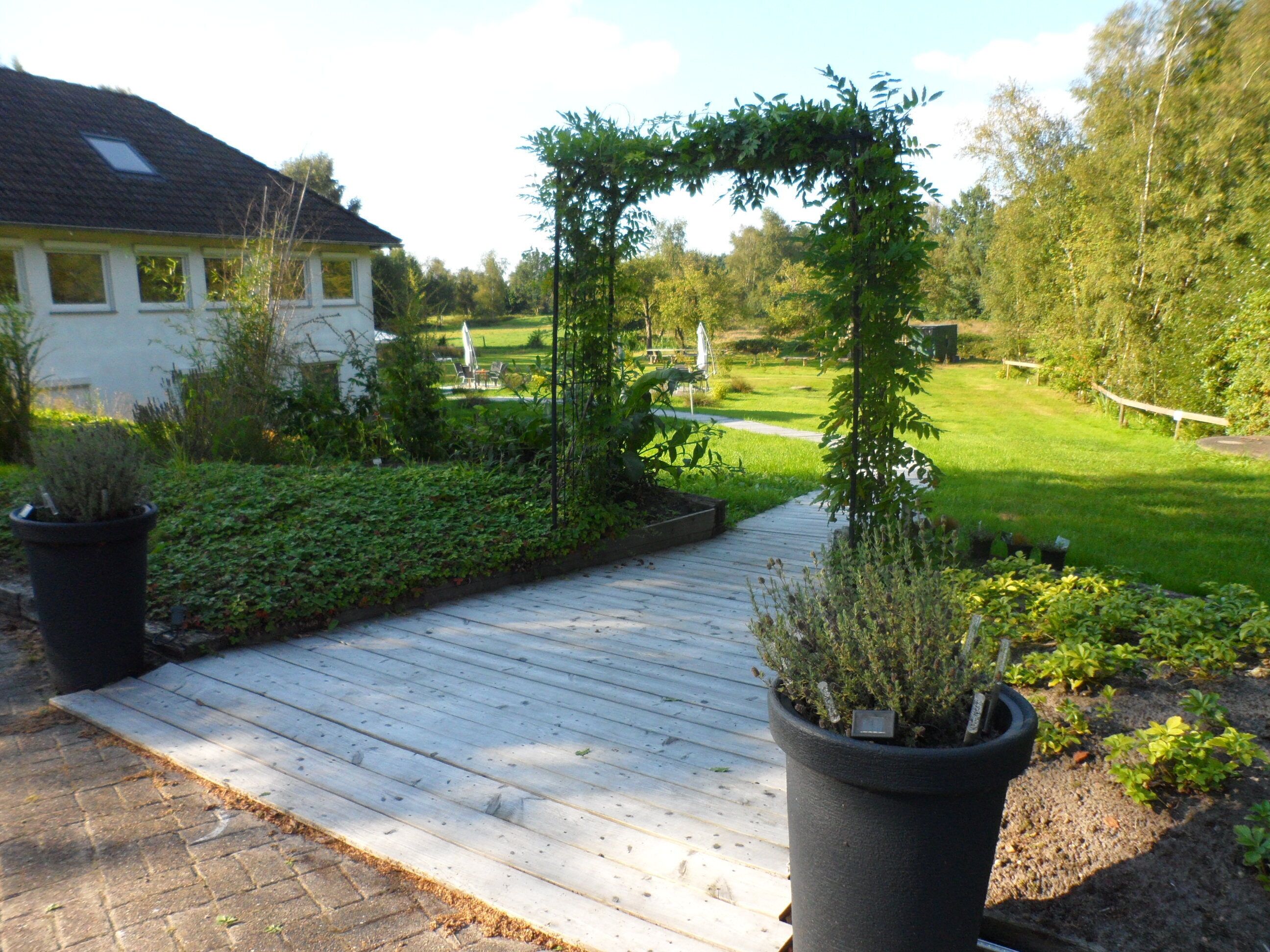 A wooden deck leading to a garden area with potted plants and a white house in the background.
