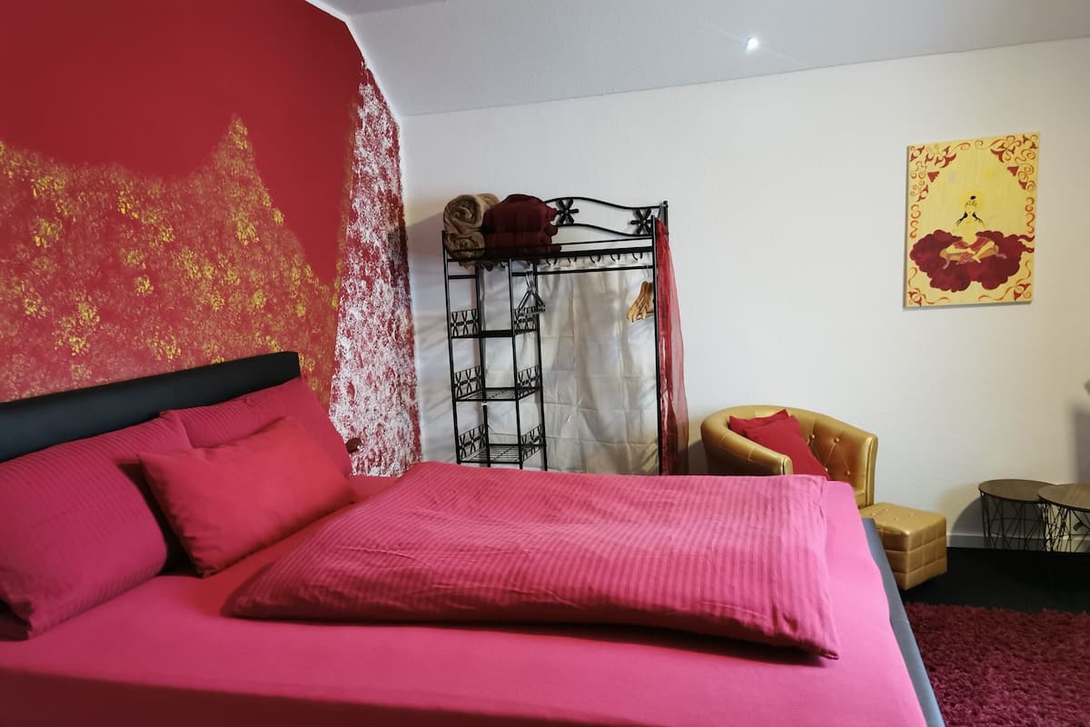 A bed with a red bedspread and pillows, a black metal headboard, a red curtain, a gold chair, and a yellow artwork on the wall.