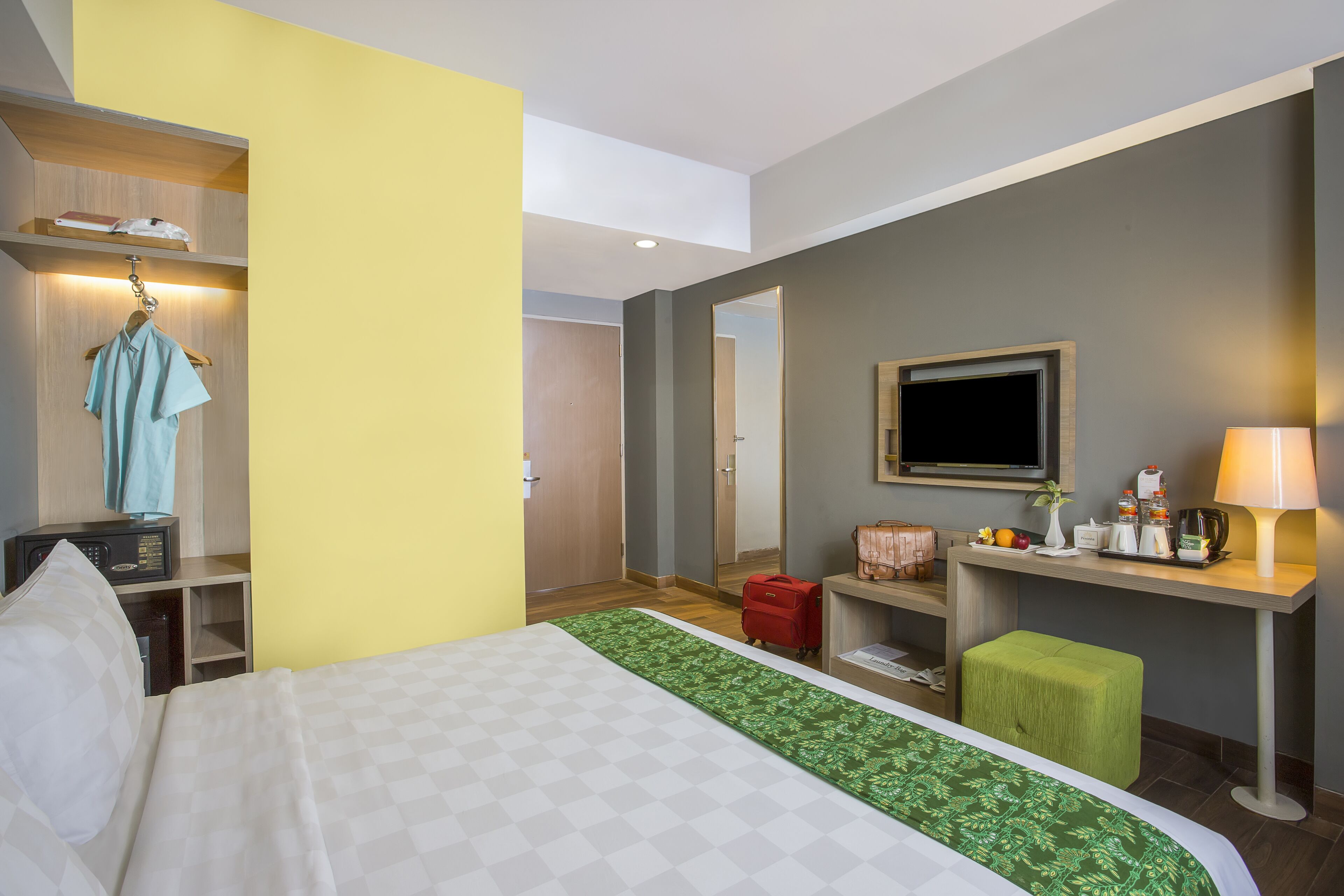 A hotel room with a bed, a desk with a lamp, a television, and a small green stool.