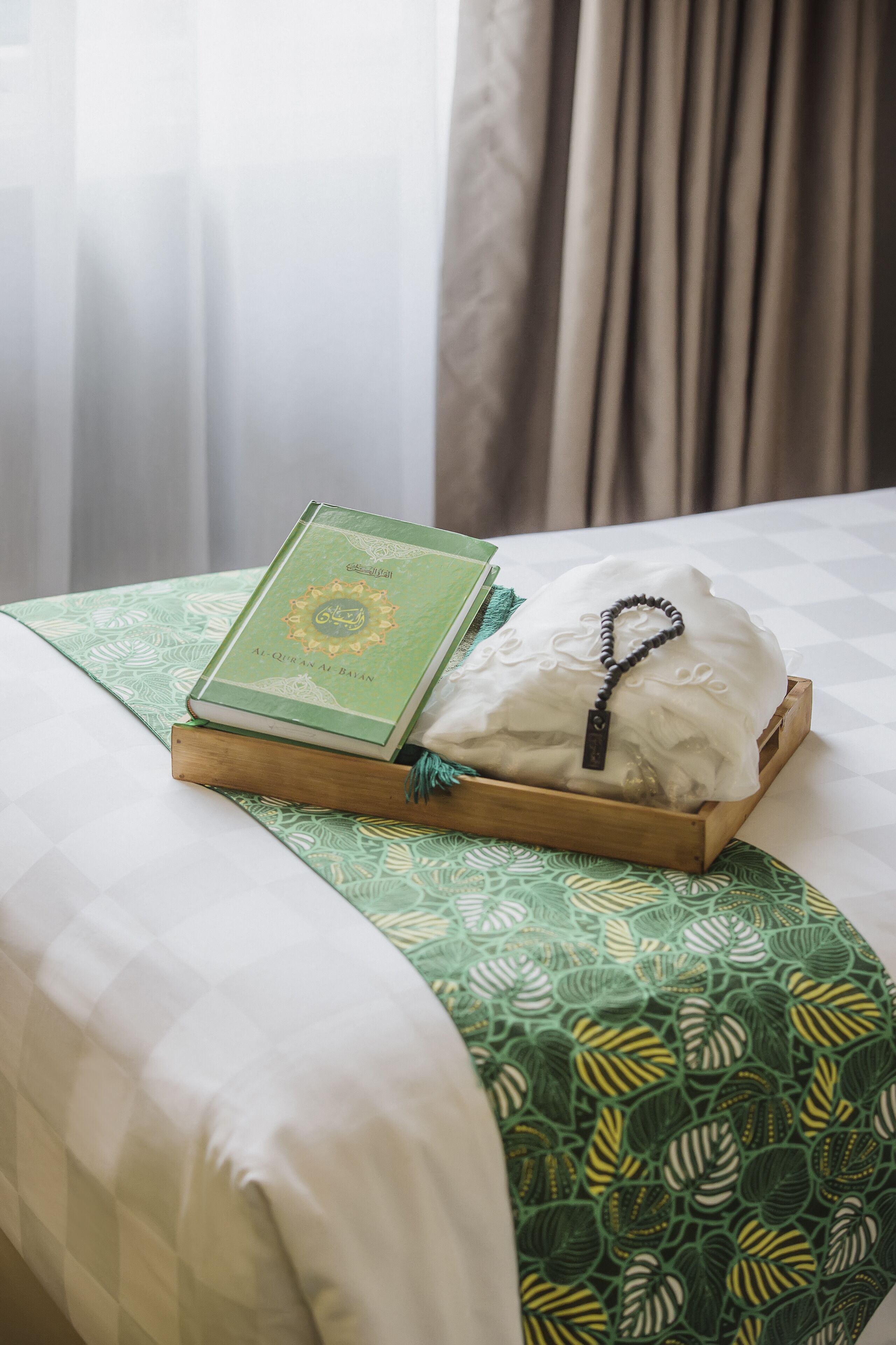 A bed with a green patterned bedspread, a book titled 'Al-Quran Al-Eemaan' on a wooden tray, and a white cloth with a black beaded necklace.