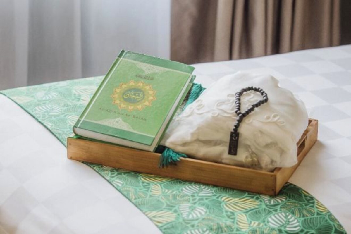 Una cama con una colcha verde estampada, una mesita de noche de madera con un libro y un paño blanco con un cordón negro.
